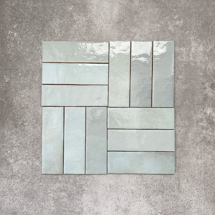 Flat lay of Zellige Aqua 6.5×20cm Moroccan-style ceramic brick tiles, shown in a basketweave layout, showcasing soft blue-green tones, a glossy handmade-look glaze and subtle tonal variation on a neutral background.