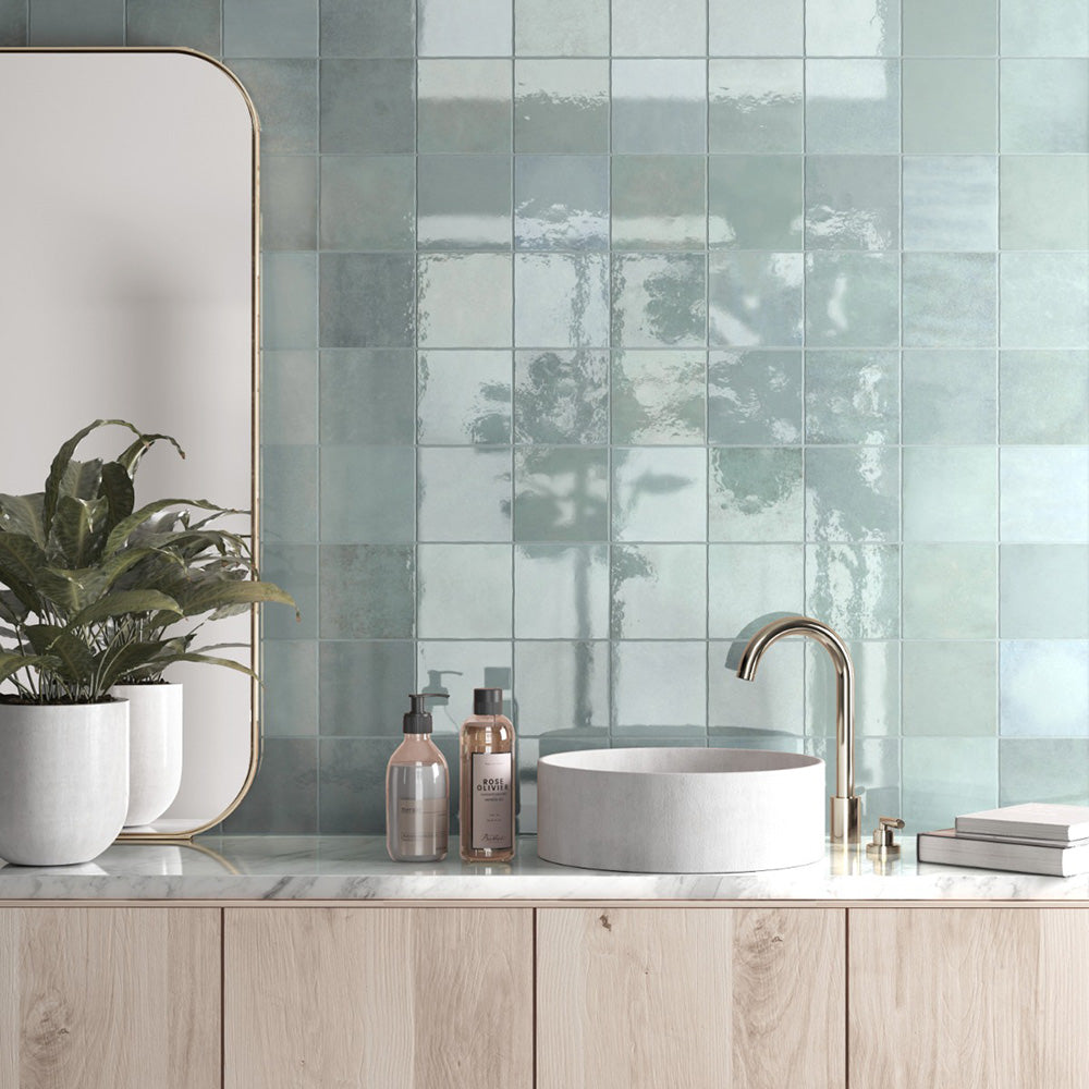 Bathroom featuring Zellige Aqua 13.2×13.2cm Moroccan-style ceramic square tiles installed on the walls behind a vanity, showcasing glossy blue-green tones with natural tonal variation, paired with a marble-effect countertop, light wood cabinetry and brushed brass tapware.
