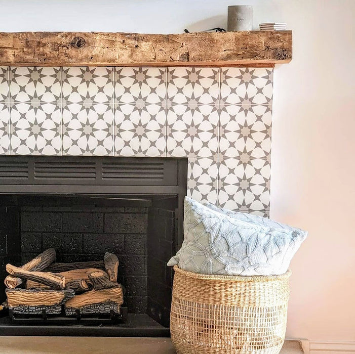 Agadir Silver Moroccan-style 20x20cm porcelain tiles used to frame a fireplace, featuring a soft grey geometric pattern paired with a rustic wooden mantel and neutral accessories for a warm, contemporary living space.