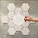 Hand holding Medina Hormuz Snow White Moroccan-style hexagon porcelain tile, 18.7 × 21.6 cm, shown against a grouped layout to highlight the crisp star-inspired geometric pattern in white and soft grey tones.