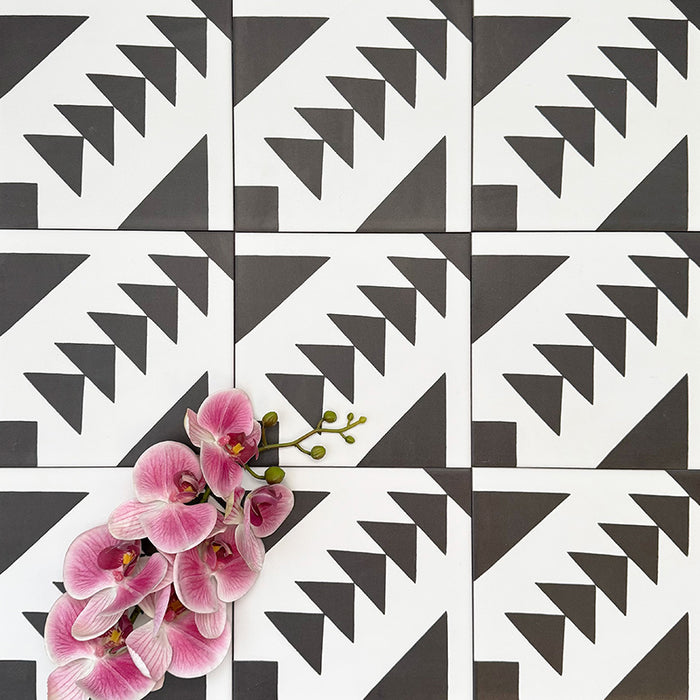 Almeira Azteca Moroccan-style porcelain tiles arranged in a repeating pattern, highlighting the bold black and white geometric design with stepped triangles, styled with pink flowers for contrast, 20x20cm.