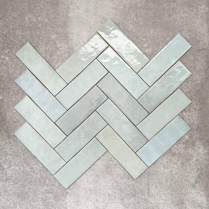 Flat lay of Zellige Aqua 6.5×20cm Moroccan-style ceramic brick tiles, shown in a herringbone layout, showcasing soft blue-green tones, a glossy handmade-look glaze and subtle tonal variation on a neutral background.