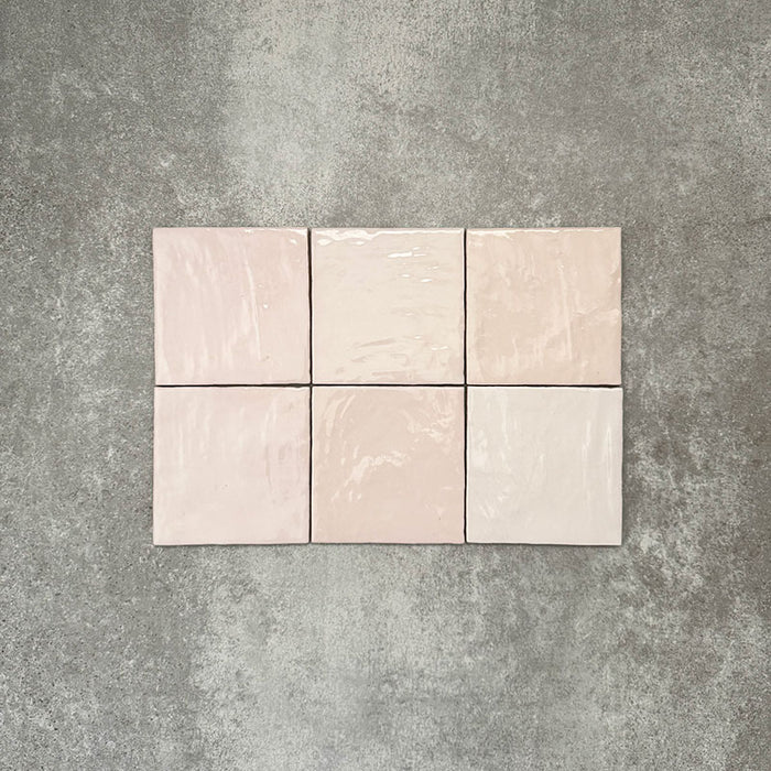 Flat lay of Zellige Pink Mist Square 13.2 × 13.2cm Moroccan-style ceramic tiles in soft pale pink and blush tones, arranged in a simple grid on a textured grey background, showcasing the glossy glaze, subtle tonal variation and handcrafted zellige-effect finish.