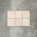Flat lay of Zellige Pink Mist Square 13.2 × 13.2cm Moroccan-style ceramic tiles in soft pale pink and blush tones, arranged in a simple grid on a textured grey background, showcasing the glossy glaze, subtle tonal variation and handcrafted zellige-effect finish.